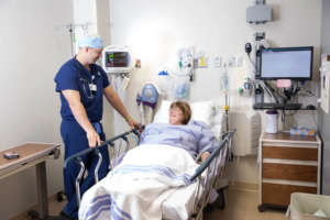 A male CRNA speaks with a female patient at her bedside.