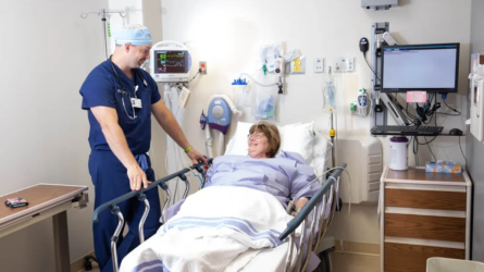 A male CRNA speaks with a female patient at her bedside.